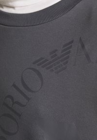 Sudadera gris oscuro con cuello acanalado, que presenta un gran diseño de logo en relieve, tejido texturizado y sutiles acentos de marca.