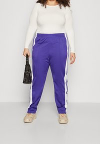 Fialové tepláky s bílými bočními pruhy, s elastickým pasem. V kombinaci s přiléhavým bílým topem s dlouhým rukávem a béžovou obuví.