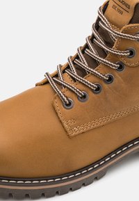 Bota de cuero marrón con acentos en tonos marrón, que presenta ojales de metal, cordones gruesos en un patrón de zigzag y una suela de goma negra resistente.