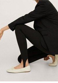 Personne accroupie portant des leggings noirs avec des fentes aux chevilles, un blazer noir et des ballerines crème ornées de petits nœuds.