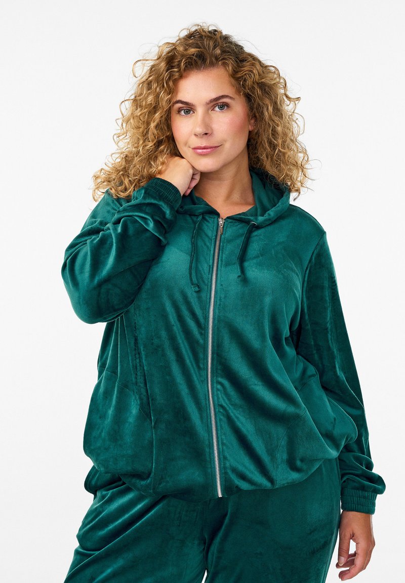 Felpa con zip in velluto verde scuro, dal fit rilassato, con ampie tasche frontali e cappuccio regolabile con lacci. Texture morbida con polsini a costine.