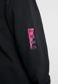 Manica nera con un patch ricamato rosa e nero che mostra le lettere "GRWY" in filo viola e nero.