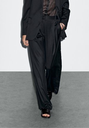 Persoon die losse zwarte broek, een zwart colbert, doorzichtige zwarte top en zwarte open teen sandalen met hak draagt, lopend op een grijze vloer.