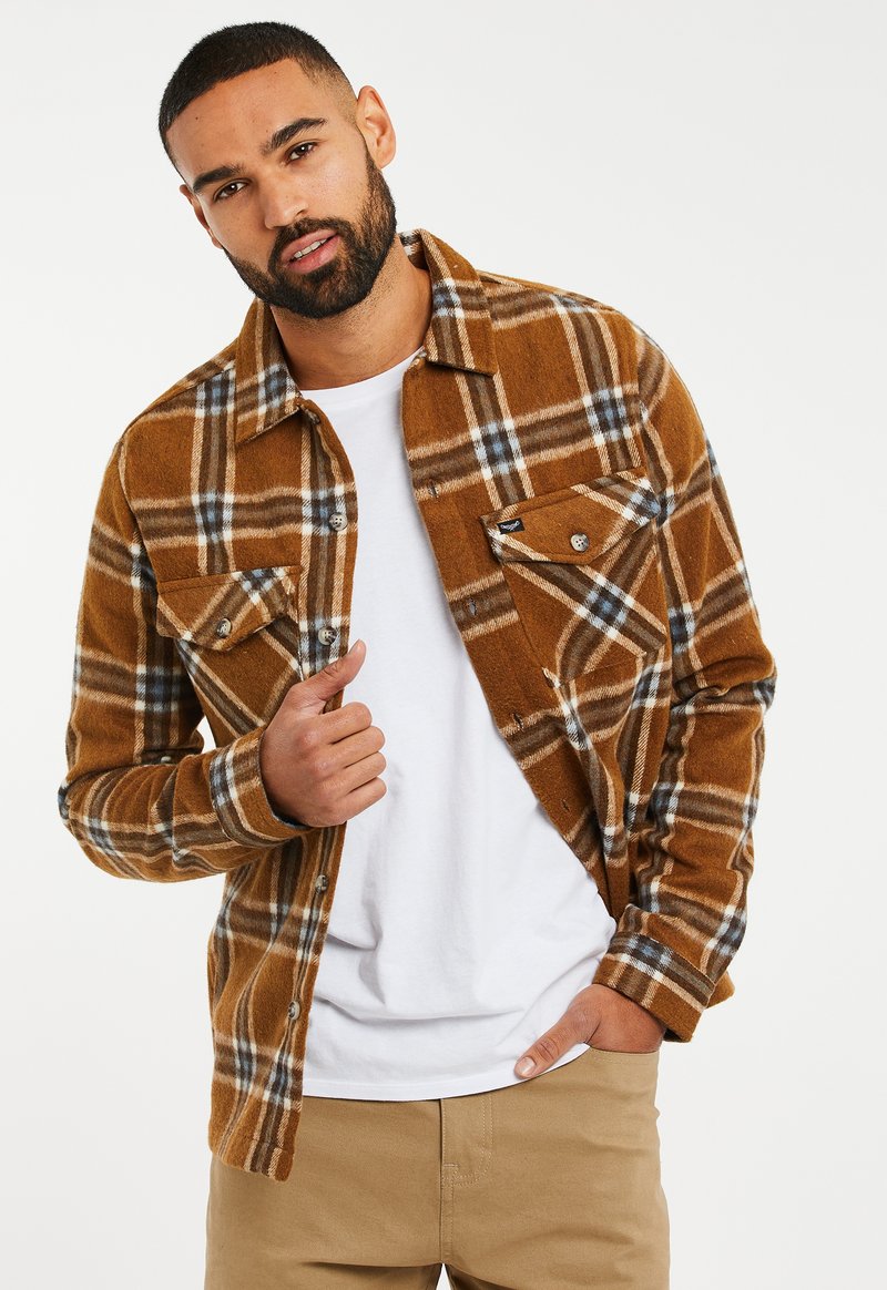 Brown plaid flannel shirt with white and blue lines, featuring snap-button chest pockets and a relaxed fit, layered over a white t-shirt.