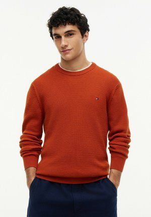Ung mand med mørkt krøllet hår iført en tekstureret orange sweater og mørkeblå bukser, hænderne i lommerne, neutralt baggrund.