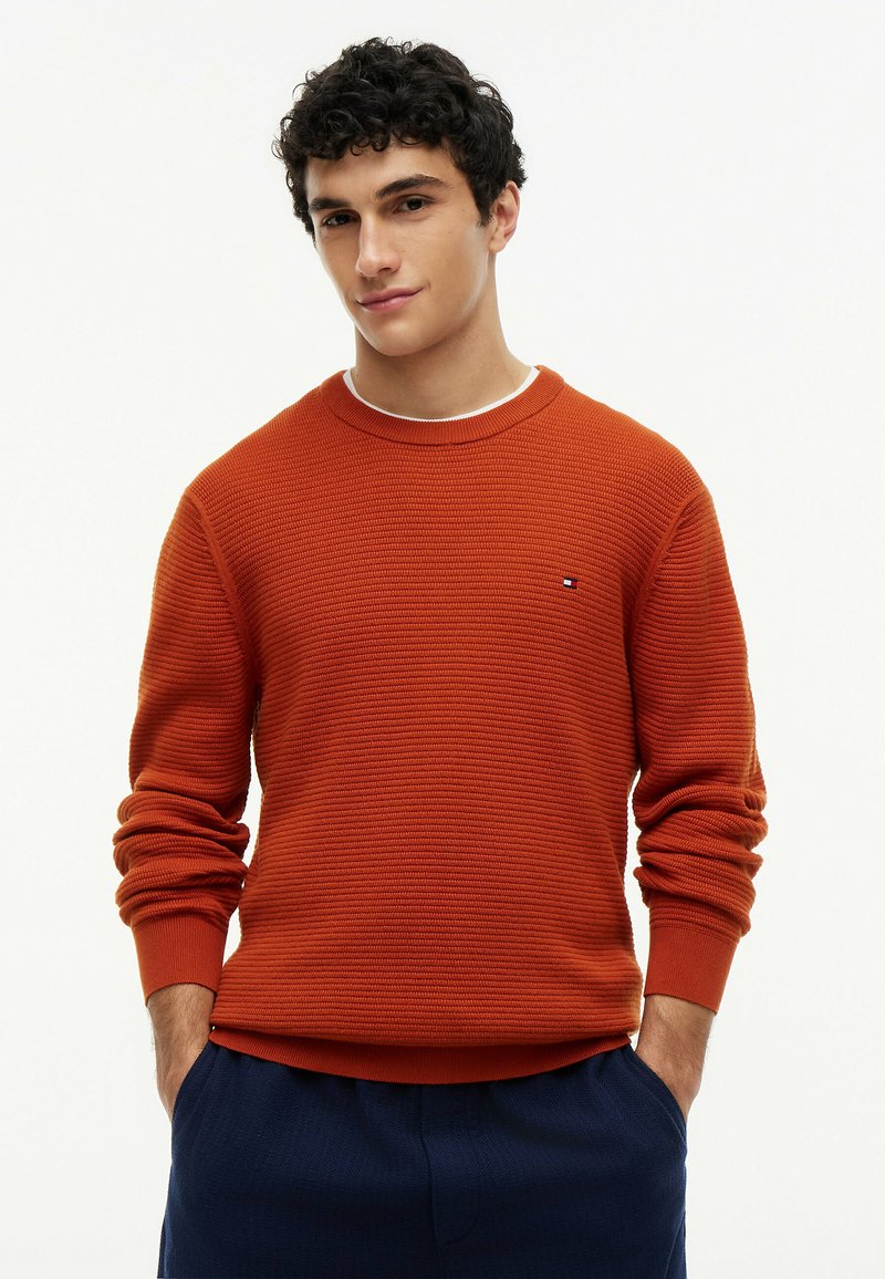 Jeune homme aux cheveux bouclés foncés portant un pull orange texturé et un pantalon bleu foncé, mains dans les poches, fond neutre.