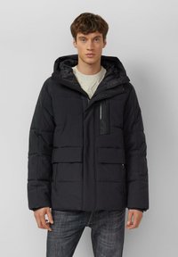 Schwarze Steppjacke mit Kapuze, mehreren Taschen, Reißverschluss und gesteppter Textur. Getragen über einem cremefarbenen Strickpullover.