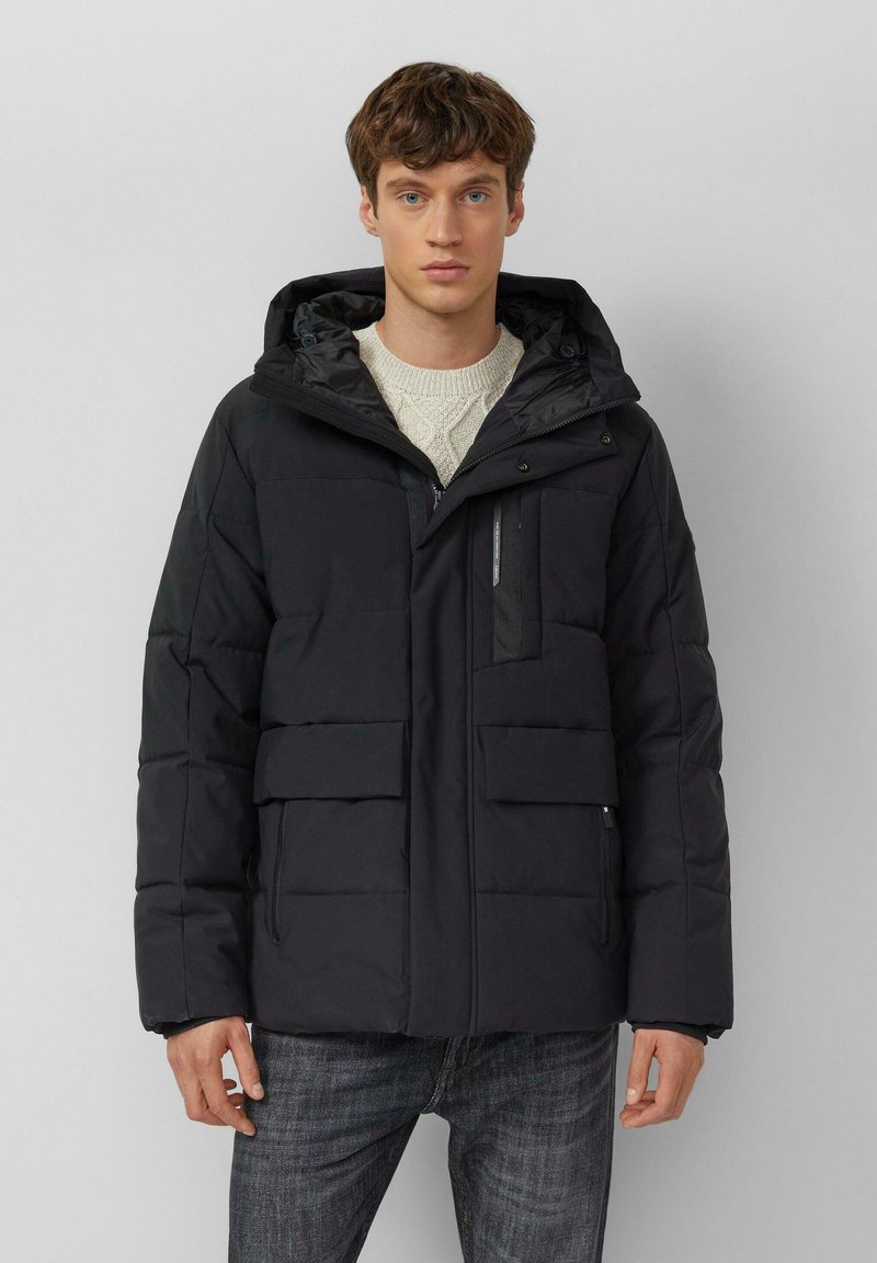 Schwarze Steppjacke mit Kapuze, mehreren Taschen, Reißverschluss und gesteppter Textur. Getragen über einem cremefarbenen Strickpullover.