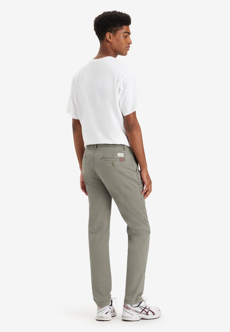Pantalon en coton gris avec poches latérales et une poche arrière, associé à un t-shirt blanc ample et des baskets blanches. Design minimaliste.