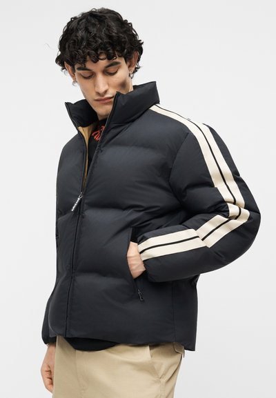 Mand iført sort dunjakke med hvide striber på ærmerne, beige bukser, ser ned med den ene hånd i jakkelommen.