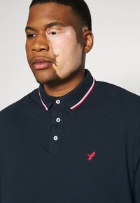 Marineblå polo med rød og hvid stripet krave, med tre knapper og lille rød brodering av en fugl på venstre bryst.