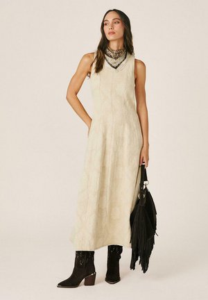 Mujer con vestido maxi beige sin mangas, pañuelo estampado en el cuello, botas negras con tachuelas y bolso negro con flecos.