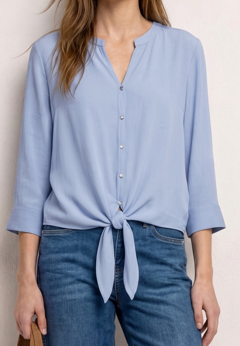 Femme portant un chemisier bleu clair boutonné avec des manches trois-quarts noué à la taille, associé à un jean bleu, tenant une anse de sac beige.