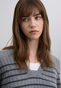 Grå ribbad tröja med V-ringning och mörkgrå horisontella ränder. Modellen har långt, rakt brunt hår och bär en vit T-shirt under.