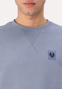 Világoskék pulóver, puha anyagból készült, kerek nyakú, elöl X-varrással díszített, és egy kis logopatch található a mellkasán.