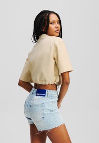 Camicetta beige tagliata con colletto rotondo, abbinata a pantaloni shorts in denim azzurro chiaro con bordi sfrangiati e patch del cinturino con marchio.