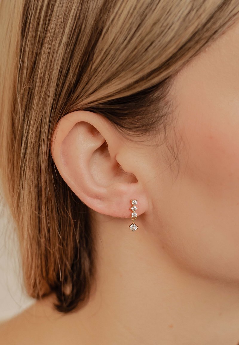 Gouden hangende oorbellen met drie heldere edelstenen in een verticale opstelling, met een bloemaccent aan de onderkant. Gladde afwerking, elegante uitstraling.