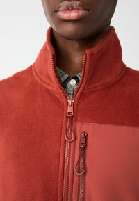 Rostfarbener Fleece-Jacke mit hohem Kragen, Reißverschluss vorne und aufgesetzter Tasche. Glatte Textur und kontrastierendes Stoffpanel.