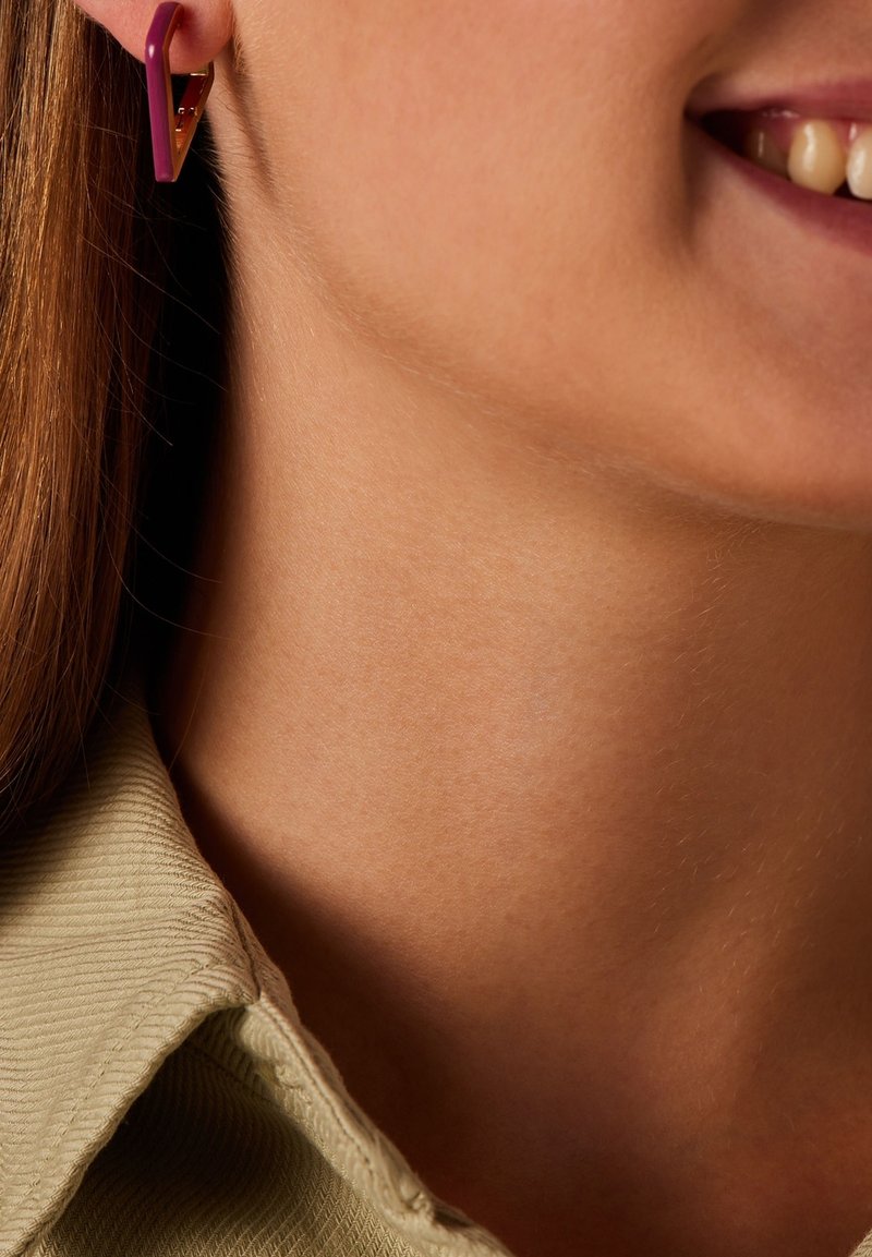 Paarse geometrische oorbel met een driehoekig ontwerp; zichtbaar gouden accent en een gladde textuur. Close-up van de nek en het haar in een lichtgroene top.