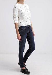Suéter blanco acanalado con lunares negros; combinado con jeans ajustados de color azul oscuro. Los zapatos son negros, planos y con cordones.