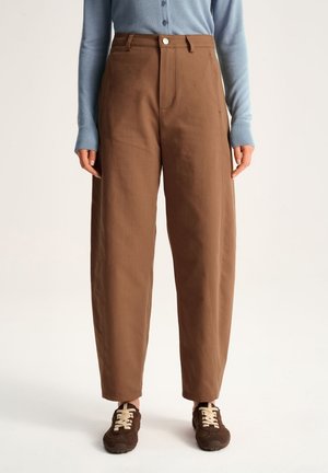 Osoba nosząca brązowe spodnie z wysokim stanem i szerokimi nogawkami, w zestawieniu z jasnoniebieskim, długim rękawem i koszulą na guziki oraz ciemnobrązowymi trampkami z beżowymi sznurowadłami.