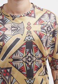 Kortärmad skjorta i beige tyg med flerfärgade geometriska mönster i röd, svart och blå, samt en rund halsdesign.