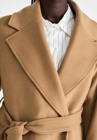 Person, der einen camel-farbenen Wollmantel mit breiten Revers und einem an der Taille gebundenen Gürtel über einem weißen Hemd mit dünnen vertikalen Streifen trägt.
