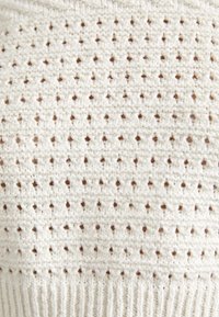 Pull en tricot avec une base crème, présentant un motif texturé de petits points bruns et un ourlet côtelé. Tissu léger et respirant.