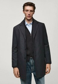 Dunkelgrauer Wollmantel mit maßgeschneiderter Passform, einreihigen Knöpfen, über einer dunklen Weste und hellblauen Hemd, kombiniert mit Jeans.