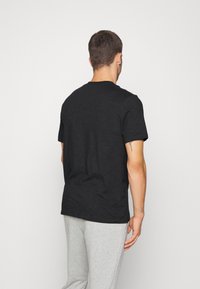 Man som vänder ryggen mot oss iförd en enkel svart t-shirt med kort ärm och ljusgråa sweatpants mot en vit bakgrund.