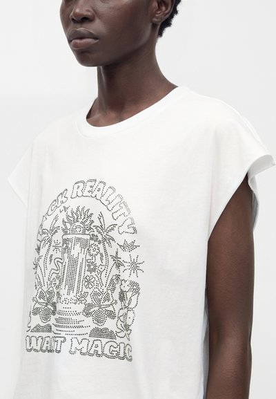 Gros plan d'une personne portant un haut blanc sans manches avec un motif floral et en colonne composé de points noirs et le texte « Fake Reality Wait Magic. »