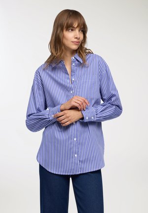 Donna che indossa una camicia a righe verticali blu e bianche con bottoni e pantaloni blu scuri, mentre aggiusta una manica su uno sfondo bianco neutro.