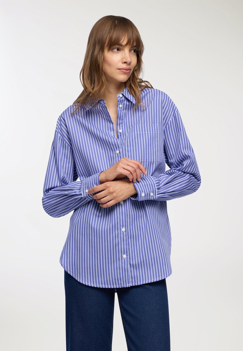 Donna che indossa una camicia a righe verticali blu e bianche con bottoni e pantaloni blu scuri, mentre aggiusta una manica su uno sfondo bianco neutro.