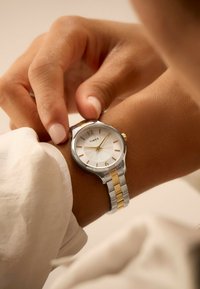 Sølv- og guldtonet armbåndsur med en tekstureret hvid urskive, timeindikatorer og et to-tone metalarmbånd. Hånden justerer uret på håndleddet.