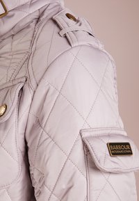 Jasnoszara kurtka pikowana z ukośnym przeszyciem, złotymi elementami oraz wyraźnym logo Barbour International na kieszeni na rękawie.