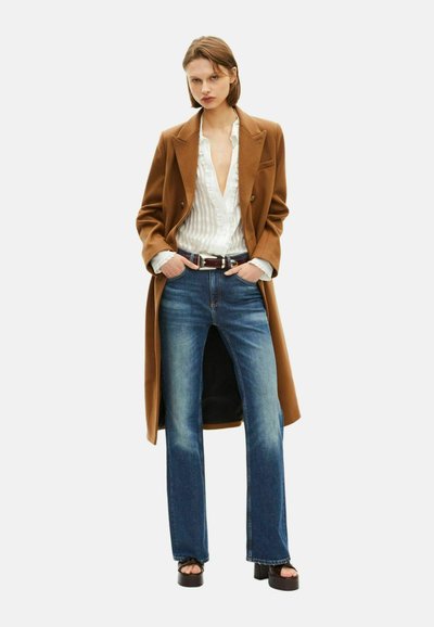 Bruine lange jas over een wit gestreept shirt, gecombineerd met blauwe uitlopende jeans en zwarte platform sandalen. De outfit bevat een bruine riem.