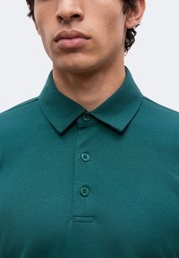 Jeune homme portant un polo vert à col et trois boutons, montrant le bas du visage et le cou sur un fond uni.