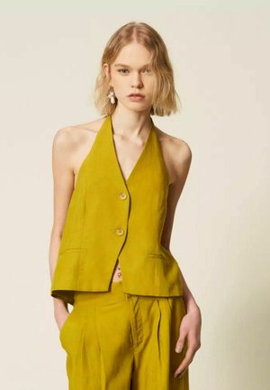 Giovane donna con capelli biondi corti che indossa un gilet smanicato giallo senape e pantaloni a vita alta abbinati, in piedi davanti a uno sfondo neutro.