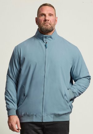 Mand med skæg iført lyseblå lynlåstrøje med knappet krave og hænderne i lommerne, vendt mod fronten mod en ensfarvet baggrund.