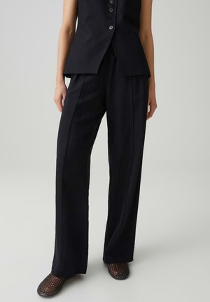 Persoon die zwarte broek met wijde pijpen, een mouwloos zwart topje met knopen en zwarte, gevlochten instapschoenen draagt, staand tegen een lichtgrijze achtergrond.