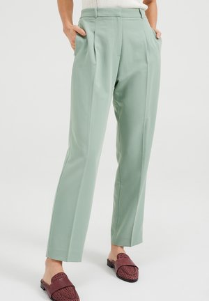 Femme portant un pantalon ajusté vert clair avec poches latérales et des chaussures plates bordeaux tissées, debout devant un fond uni.