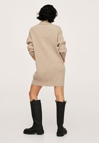 Abito a maglia beige con texture a coste e vestibilità rilassata, abbinato a stivali neri in gomma al ginocchio con suola robusta e linguette per l'estrazione.