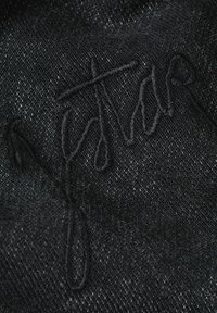 Tessuto di denim nero con una sottile texture. Presenta una firma ricamata in filo più scuro, che offre un dettaglio in rilievo sullo sfondo.