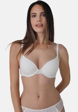 Frau mit langen dunklen Haaren, die einen weißen gepolsterten BH und passende Spitzentüllunterwäsche trägt, vor einem schlichten hellen Hintergrund stehend.