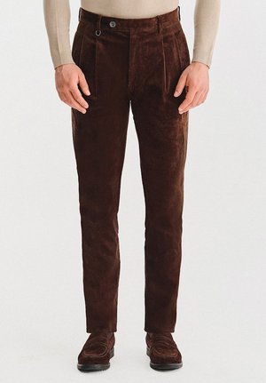 Brązowe sztruksowe spodnie o gładkiej fakturze, z dwoma plisami z przodu i zapięciem na guzik. Pasują do ciemnych butów.