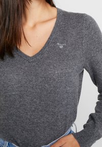 Vrouw in een grijze V-hals trui met een klein wit GANT-logo op de borst, gecombineerd met lichtblauwe jeans, hand op de heup.