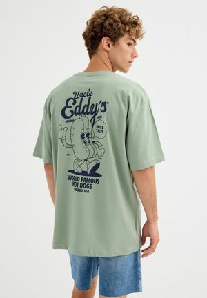 Joven con cabello rizado que lleva una camiseta grande de color verde claro con un dibujo animado de un perrito caliente y texto promocionando los perritos calientes de Tío Eddy.