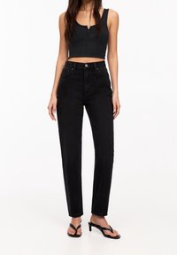 Jeans negros de cintura alta hechos de mezclilla, con corte recto, bolsillos frontales y un cierre de botón clásico. Llevados con una camiseta sin mangas negra.