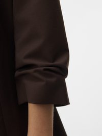 Opgerolde donkerbruine lange mouw op arm tegen een effen lichte achtergrond, met zicht op de stoftextuur en de details van de mouwmanchet.