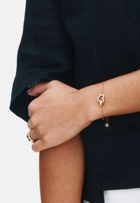 Kreisais elkonis pārkrustots pār labo apakšdelmu, valkājot melnu piedurkni, zelta gredzens uz kreisās rokas un smalka zelta aproce ar sirds rotājumu uz labā plaukstas locītavas.